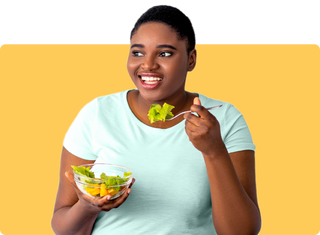 Smiling woman in a light blue t-shirt holding a glass bowl and eating a fresh salad against a yellow background