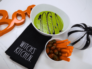 Halloween-themed table setting with green soup, carrots, and a 'Witch's Kitchen' towel.