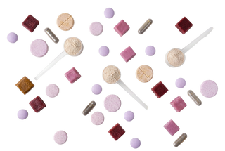 An overhead flat-lay of assorted pills and capsules scattered on a white background, including round lavender and pink tablets, dark red square tablets, beige powder scoops, and silver capsules.