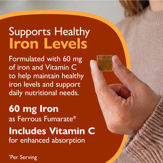 A close-up of a hand holding a small brown rectangular supplement tablet, with large text on the left stating it supports healthy iron levels and includes Vitamin C. The background is a soft beige indoor setting with part of a person's arm and sweater visible.