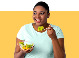 A smiling woman with short dark hair wearing a light mint green t-shirt holds a clear bowl of salad and a fork with lettuce, posed against a split white and yellow background.